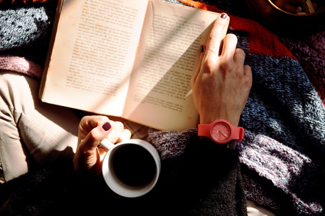 birds-eye view of a women reading book while holding a mug of black coffee