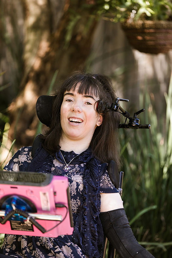 A young woman with cerebral palsy is pictured in her mobility device smiling at the camera.