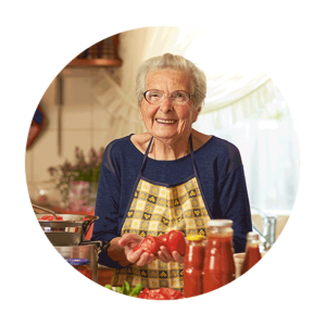 A senior women wearing glasses and and apron holds chopped tomatoes while smiling at the camera