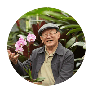 A senior man sits in a garden and holds a pink blooming orchid. He is wearing a hat and glasses and smiles at the camera